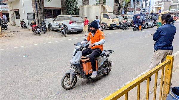 A Swiggy delivery rider in an orange uniform on an electric scooter on a city street